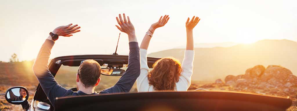 couple from behind in convertible car with hands raised, looking across the desert.