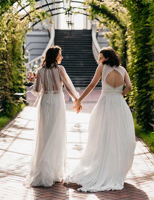 lesbian couple in wedding gowns walking down path.