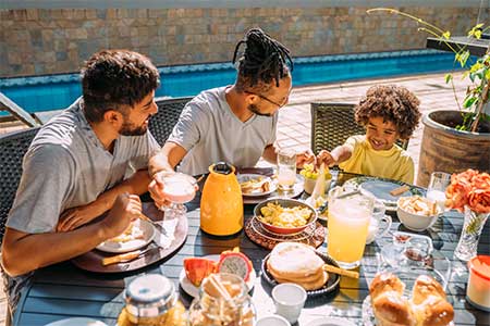 gay male couple with young child sitting at table for breakfast outdoors.