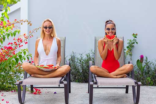 two women in bathing suits on lounge chairs eating watermelon.